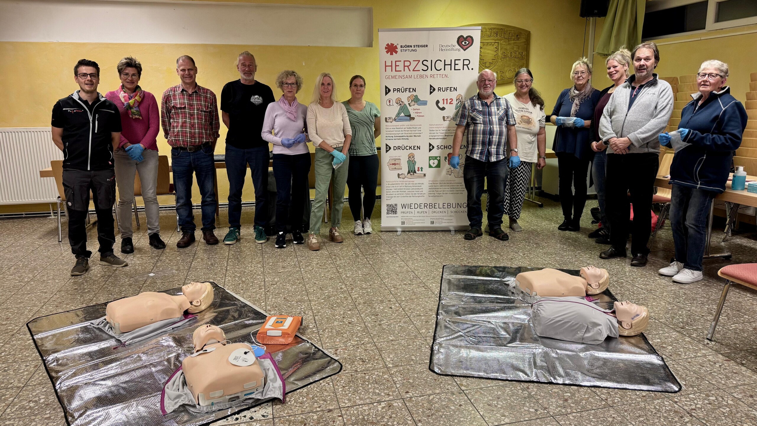 Gruppenfoto des HERZSICHER-Kurses in Goldbeck mit Teilnehmenden, Übungspuppen und AED-Trainingsgerät.