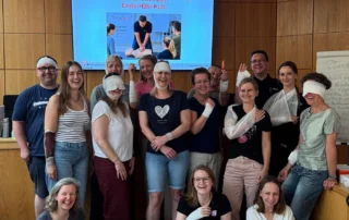Teilnehmende des Erste-Hilfe-Kurses im Amtsgericht Bielefeld mit Übungsverbänden, Reanimationspuppe und AED-Trainingsgerät.